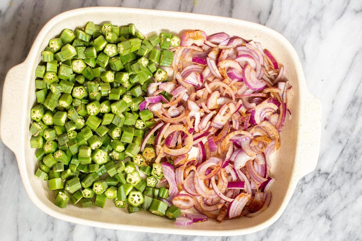 Indian spices being sprinkled over slived onion and okra in a casserole dish