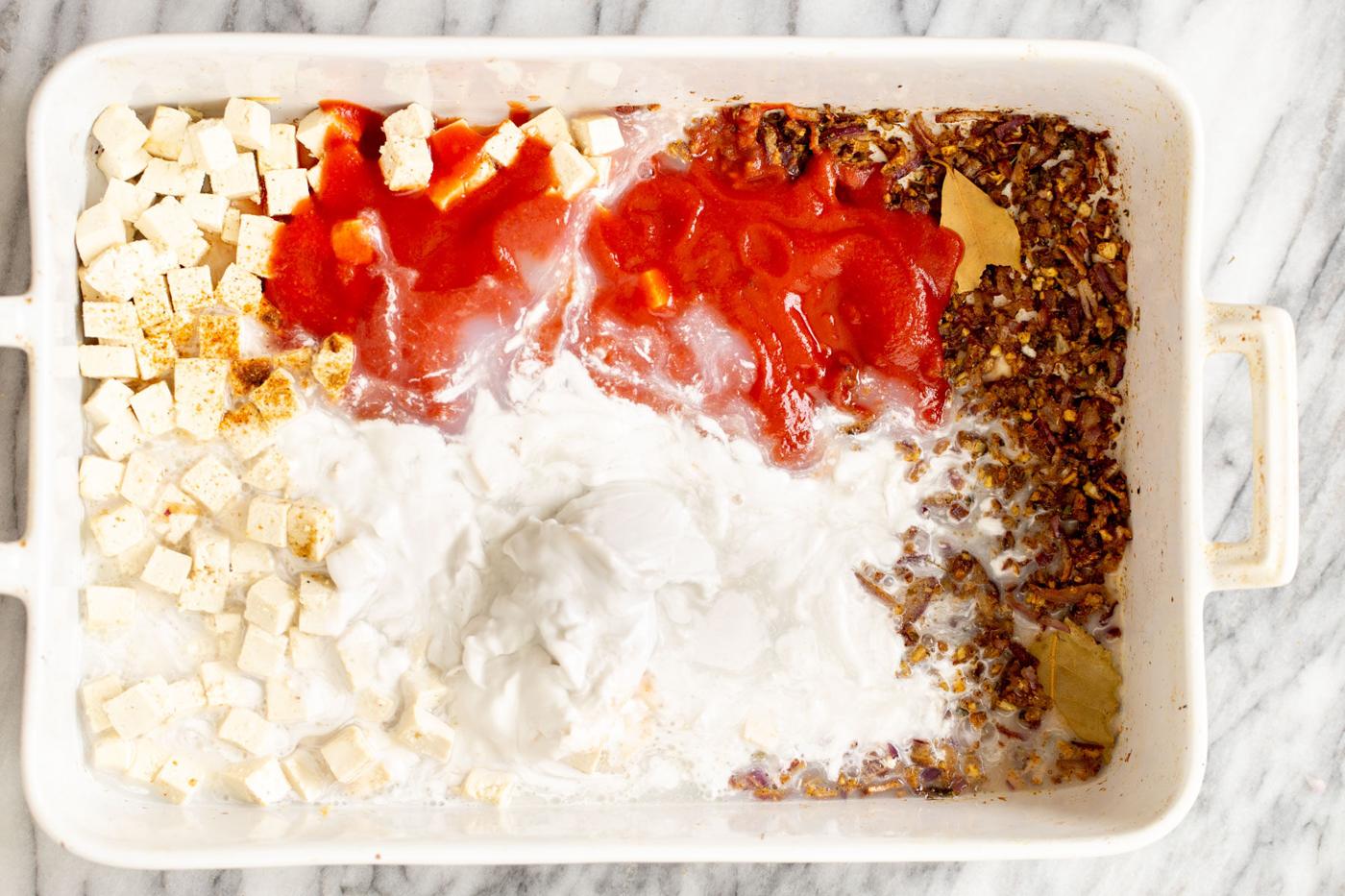 tomato puree and coconut milk being added to baked spices and tofu in casserole dish