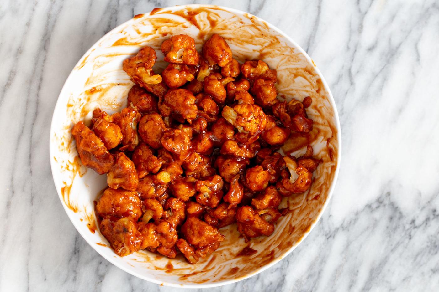 baked cauliflower wings being tossed in a small bowl with spicy bbq sauce