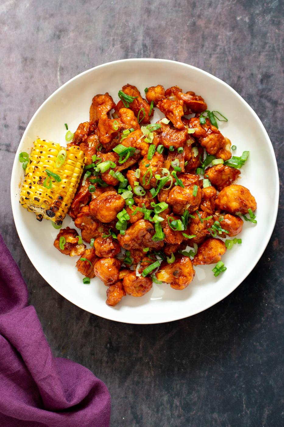 vegan baked beet battered cauliflower wings coated in bbq sauce served on a white plate with corn on the cob
