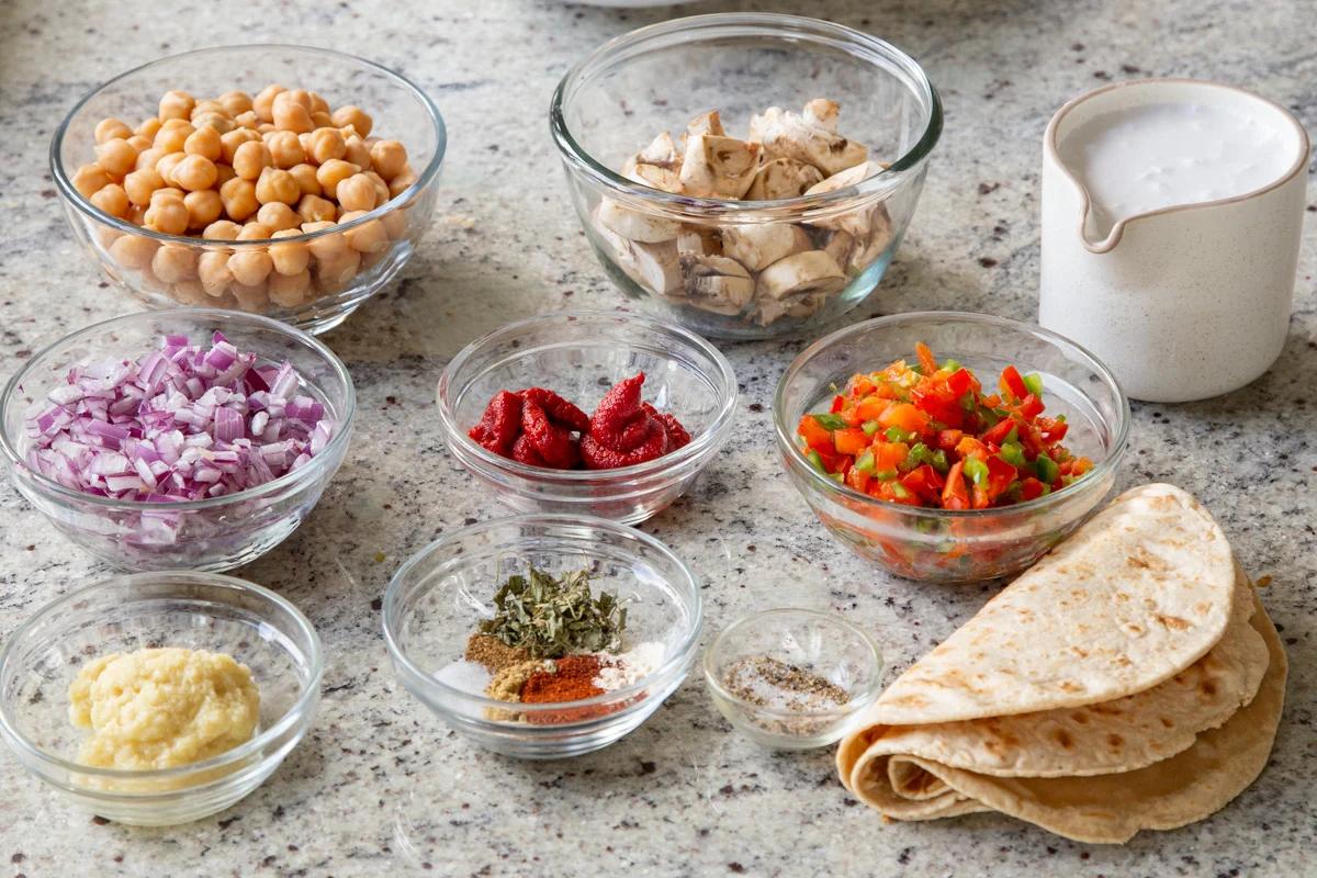 veggies, beans, tortillas, and other butter chicken enchiladas ingredients in bowls on a kitchen counter