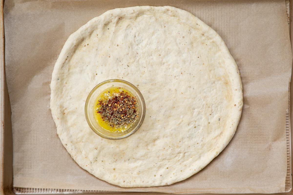 shaped unbaked vegan pizza crust with a small dish of seasoned oil on a baking sheet
