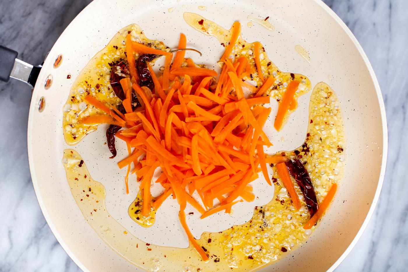 grated carrots being added to a pan with chili garlic oil