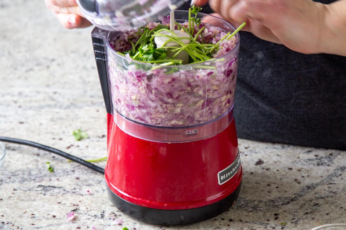 adding cilantro stems to chopped onions, garlic, ginger, and chili in a food processor
