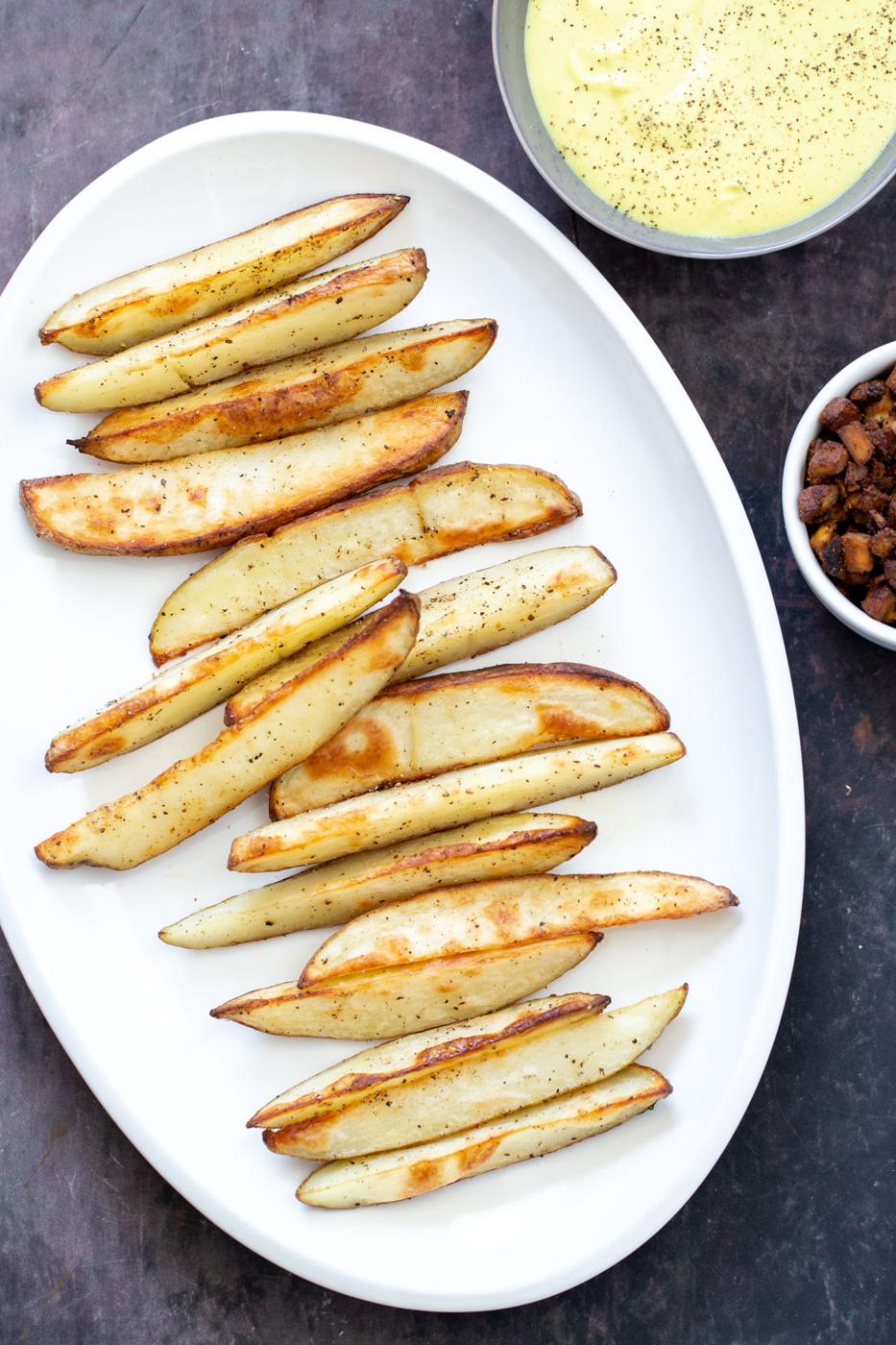 a platter of crispy vegan baked potato wedges next to a small bowl of queso and tofu bacon