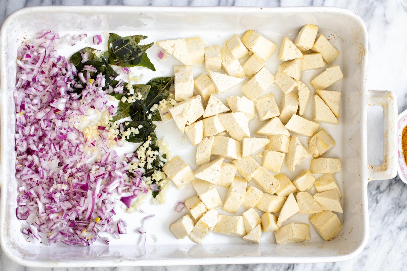 a casserole dish with cubed tofu, chopped onions and curry leaves