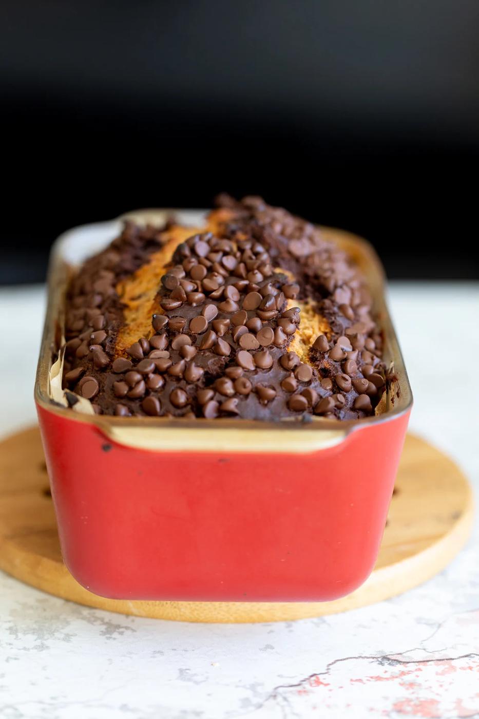 side view of a loaf of Vegan Chocolate Banana Bread topped with chocolate chips in a red ceramic loaf pan