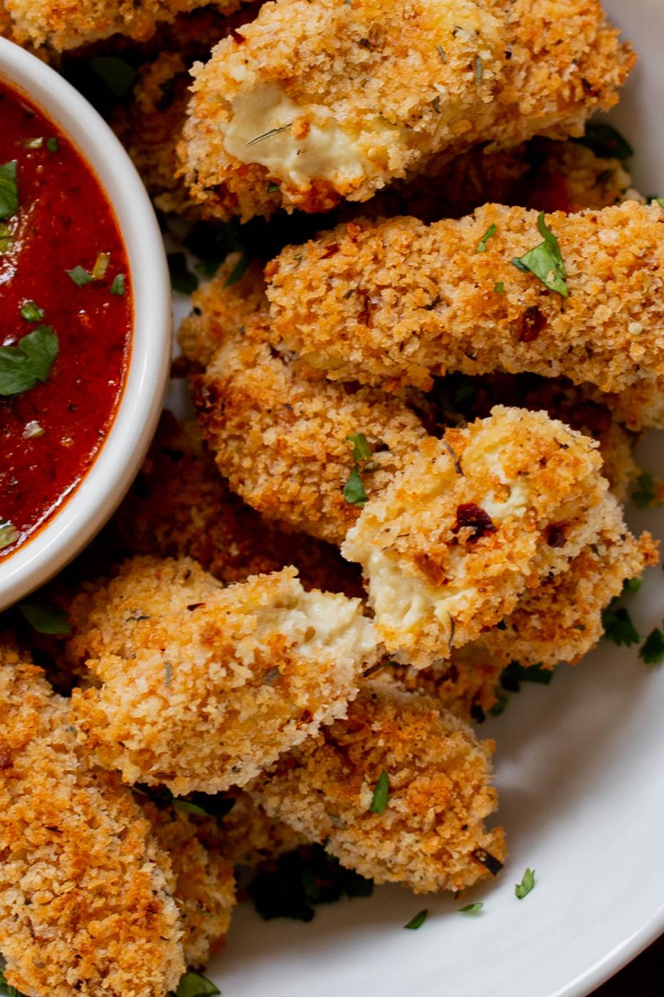 extreme close-up of vegan mozzarella sticks in a serving platter with marinara on the side with some broken sticks, so you can see the cheese