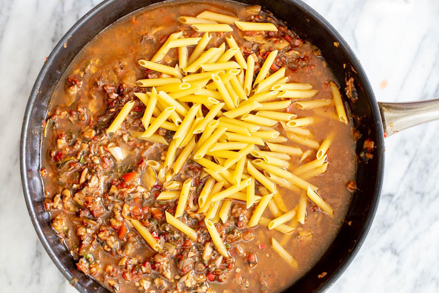 raw pasta being added to a sauteeing pan with veggies
