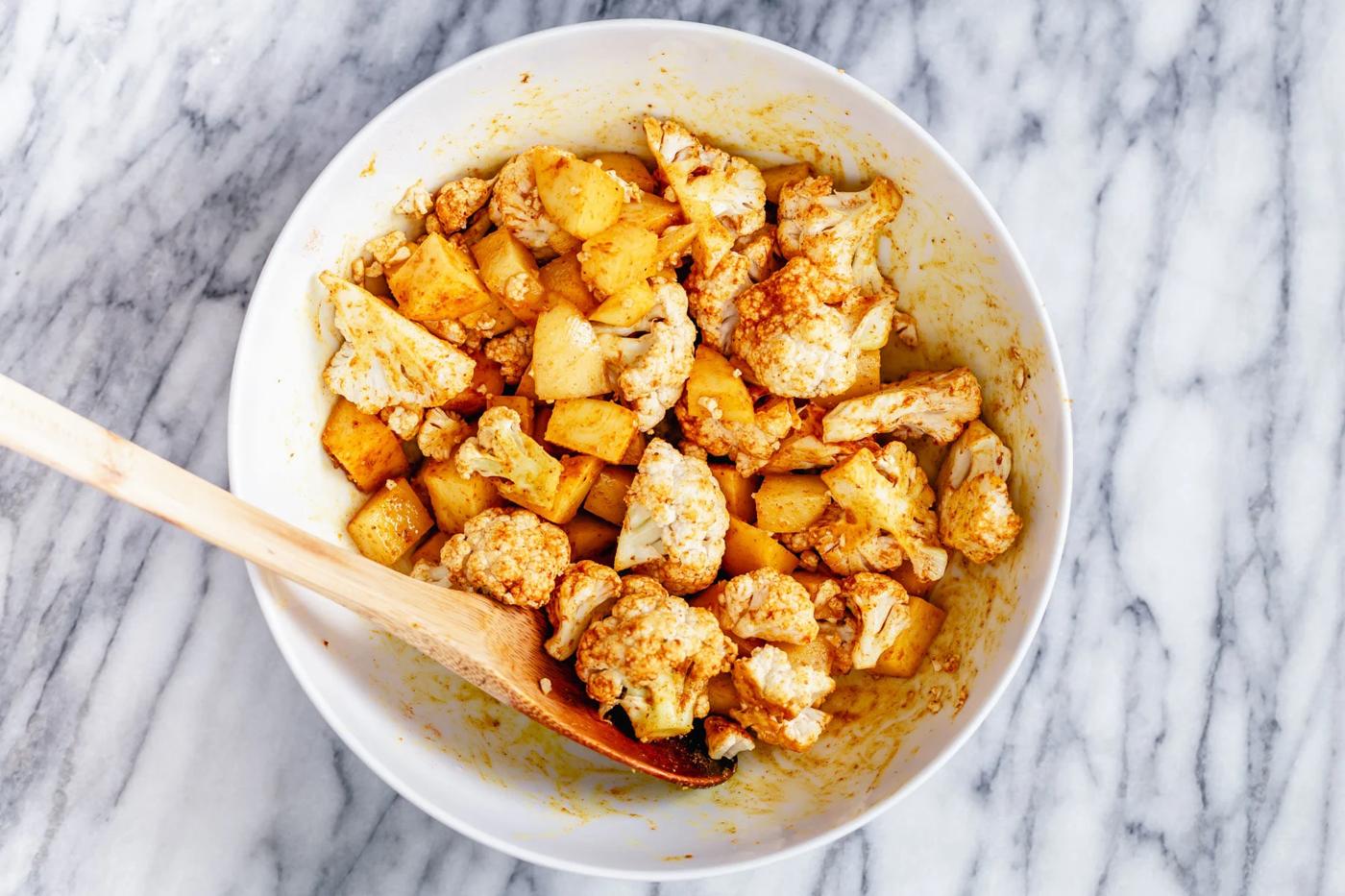 a white bowl with cauliflower and potato cubes being tossed with Indian spices