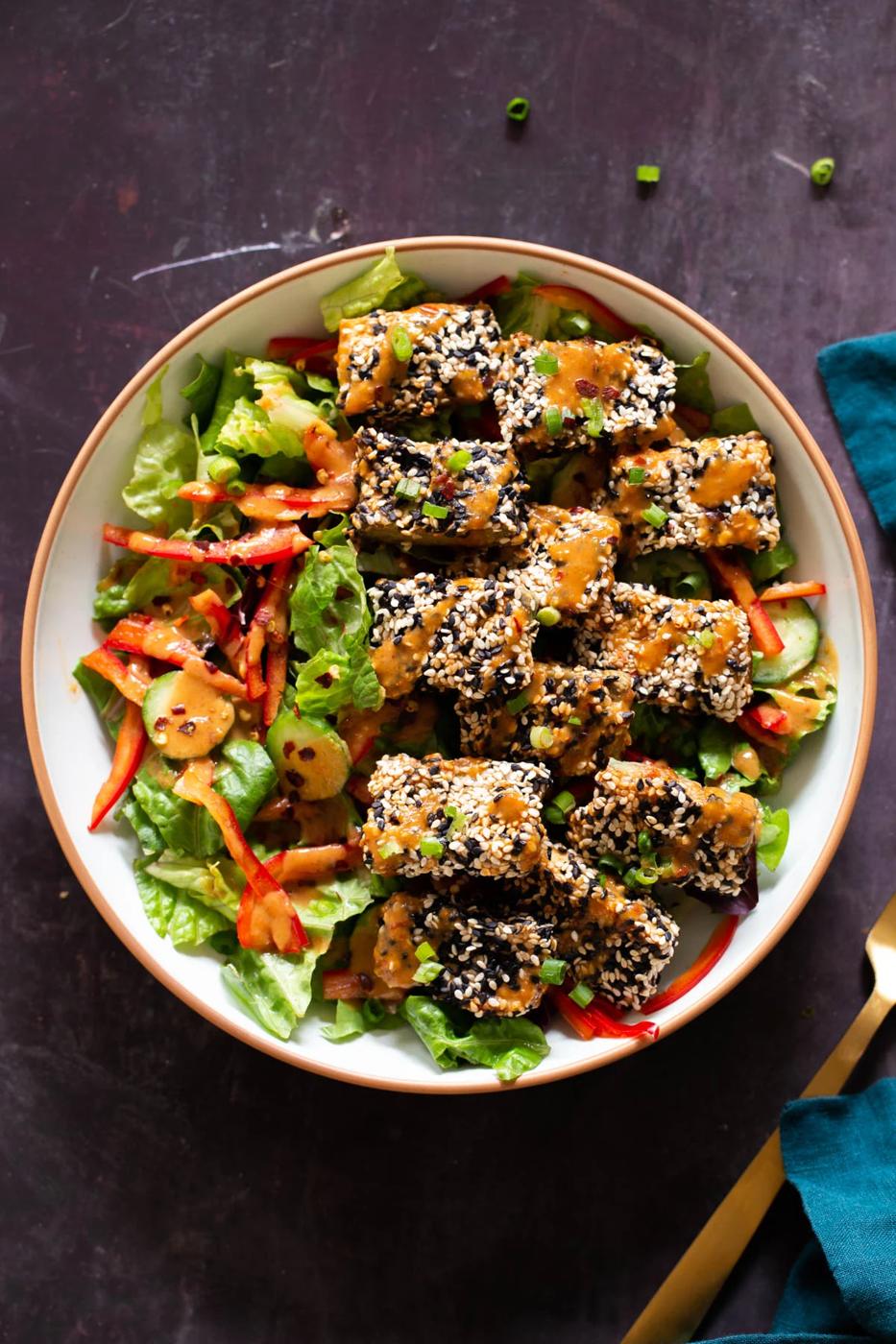 a salad bowl topped with sesame crusted tofu and drizzled with peanut sauce