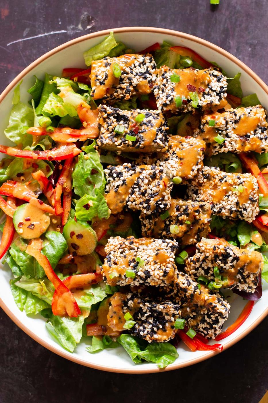 sesame crusted baked tofu on a bowl with greens and veggies drizzled with peanut sauce
