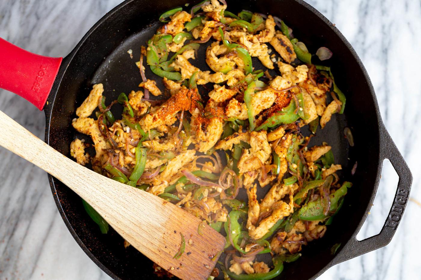 spices being added to sauteed veggies and soy curls in a black skillet