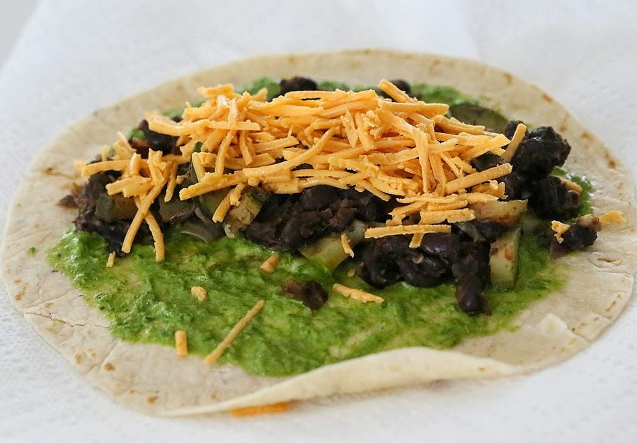a wheat wrap being topped with chimichurri, black beans and vegan shredded cheese