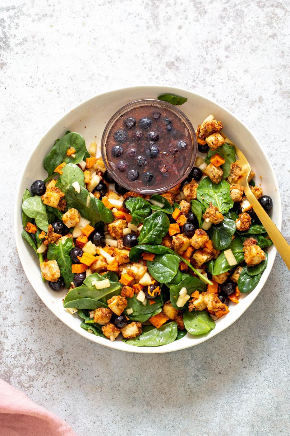 a bowl with baby spinach topped with baked sweet potatoes and crunchy tofu served with a small dish of maple blueberry dressing