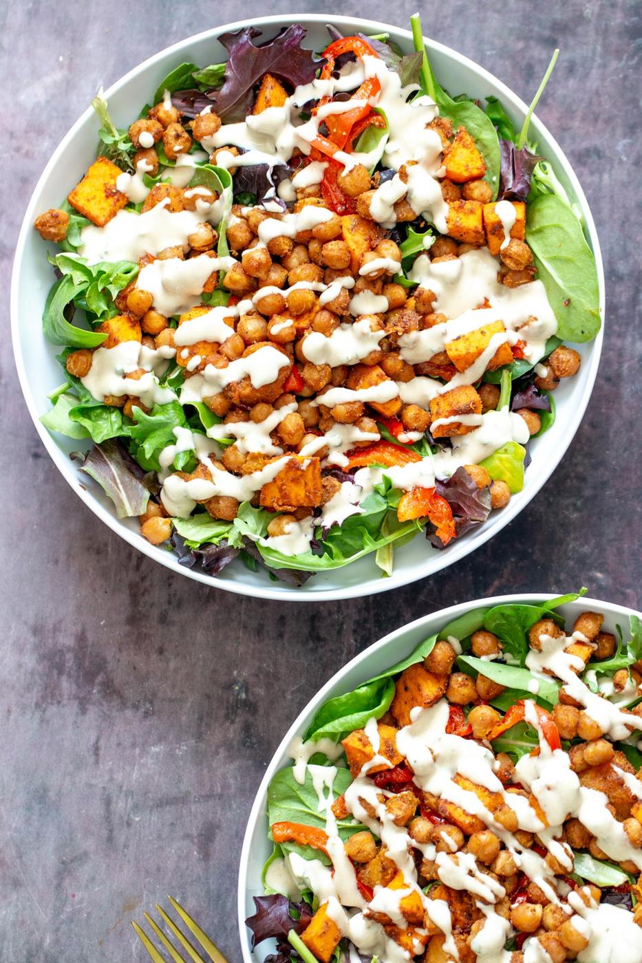 two salad bowls with spring mix topped with roasted chickpeas, bell peppers and sweet potatoes and tahini dressing