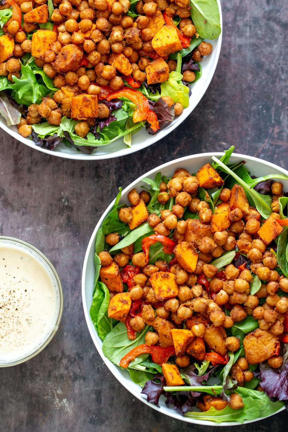 two bowls with salad topped with baked chickpeas and roasted bell pepper and sweet potatoes with a small dish of tahini dressing on the side