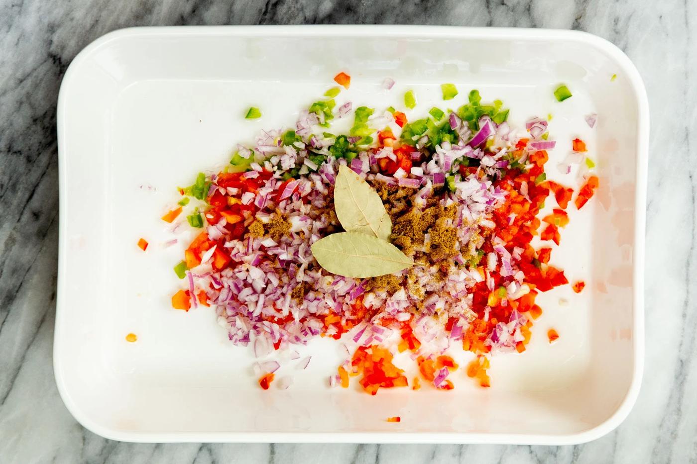 chopped bell pepper and onion with spices and bay leaves in a white casserole dish