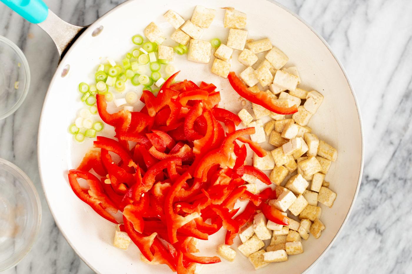 sliced bell peppers, green onion and tofu cubes being fried in a sauteeing pan