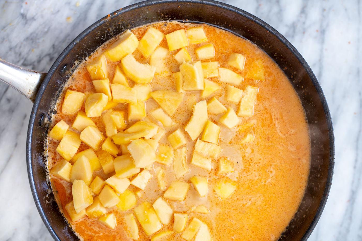 cubed fresh pumpkin being added to coconut milk broth in a saucepan