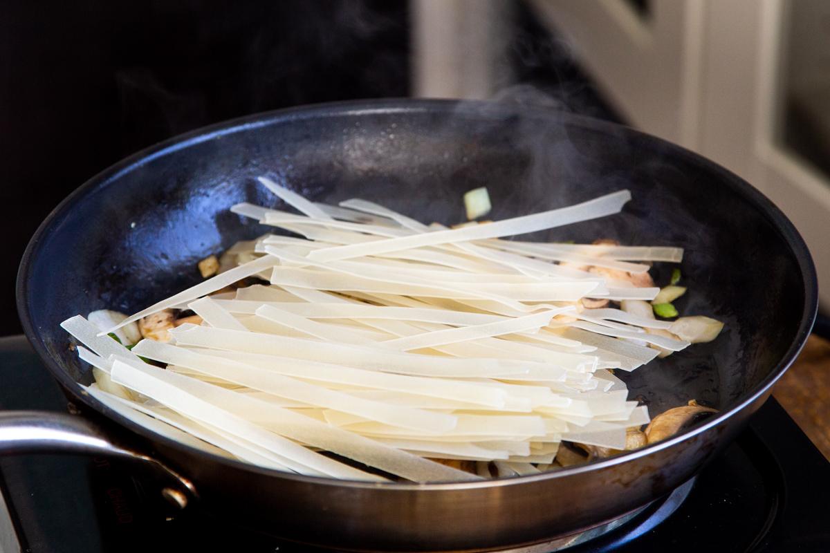 Thai rice noodles being added to a black skillet with mushrooms chili and garlic