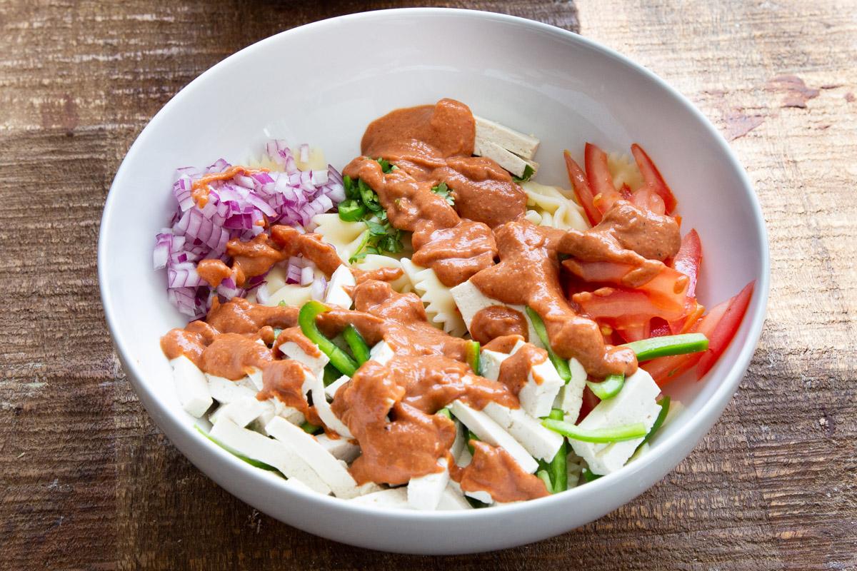 creamy tikka masala being drizzled over pasta salad ingredients in a white bowl