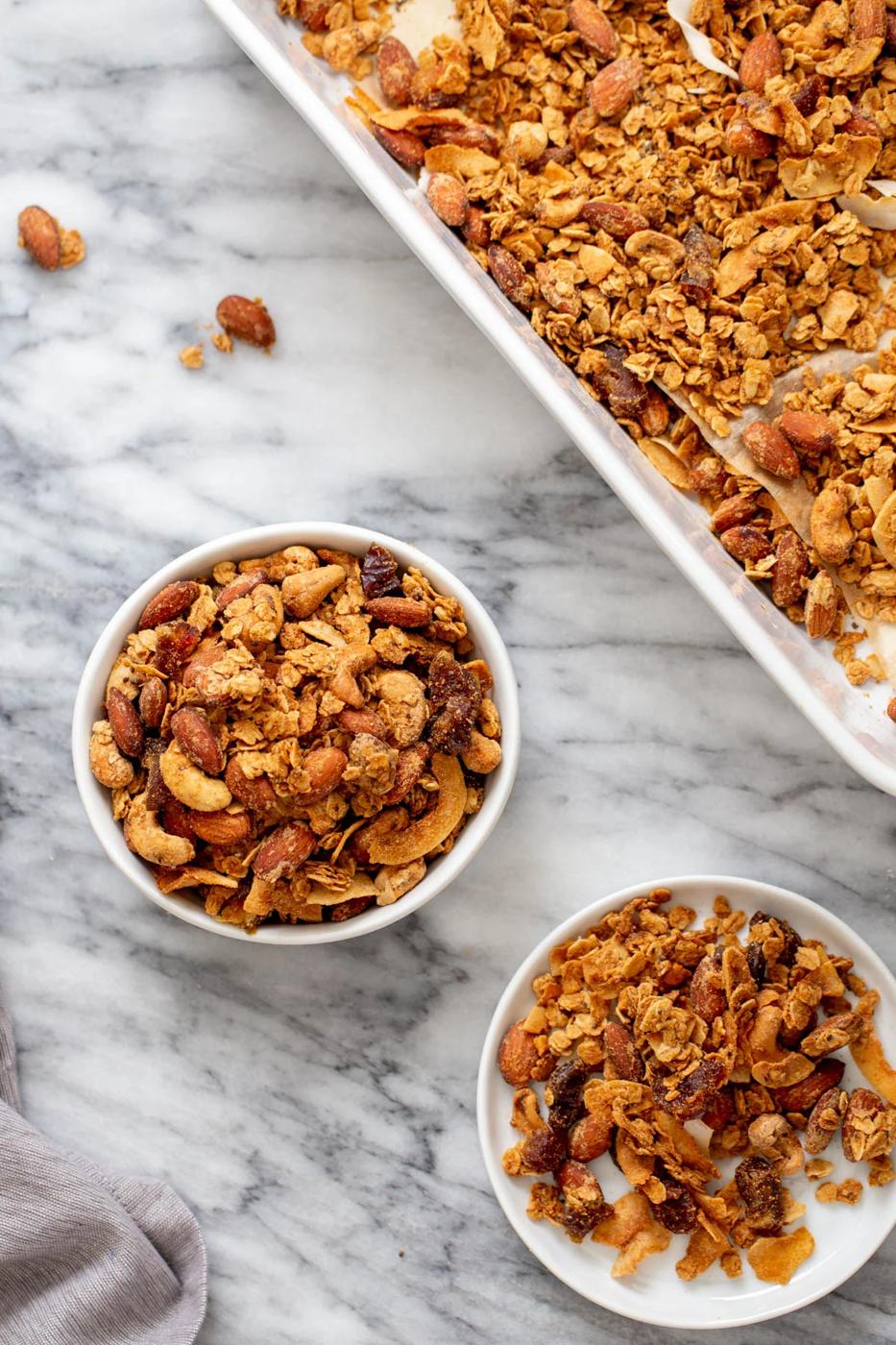 two small bowls of homemade baked vegan almond coconut granola next to a baking sheet with granola