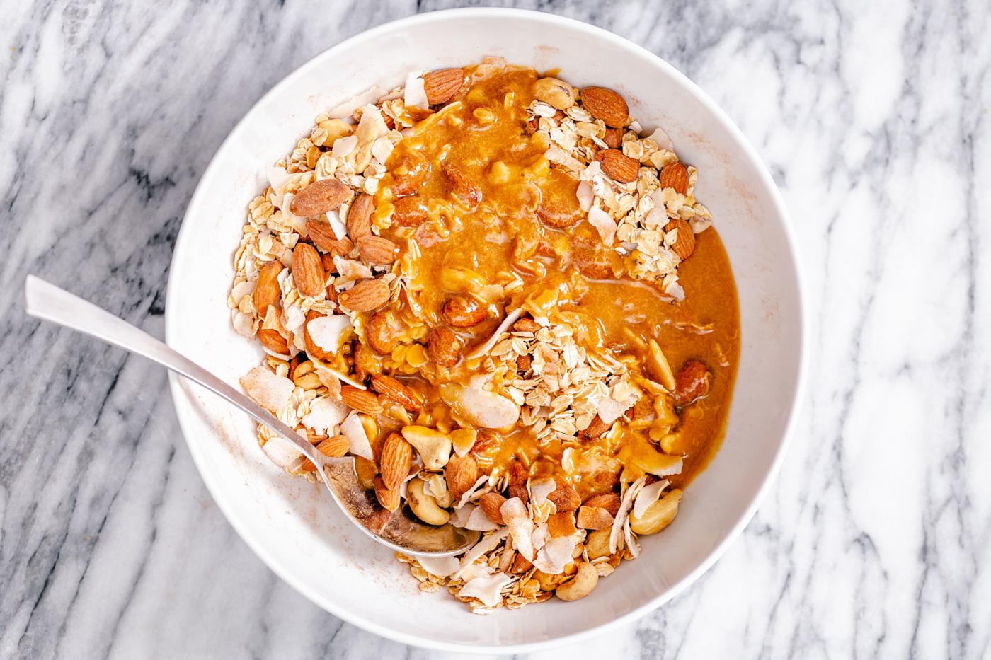 almond butter and maple syrup being added to a small bowl with dry granola mix to make baked granola