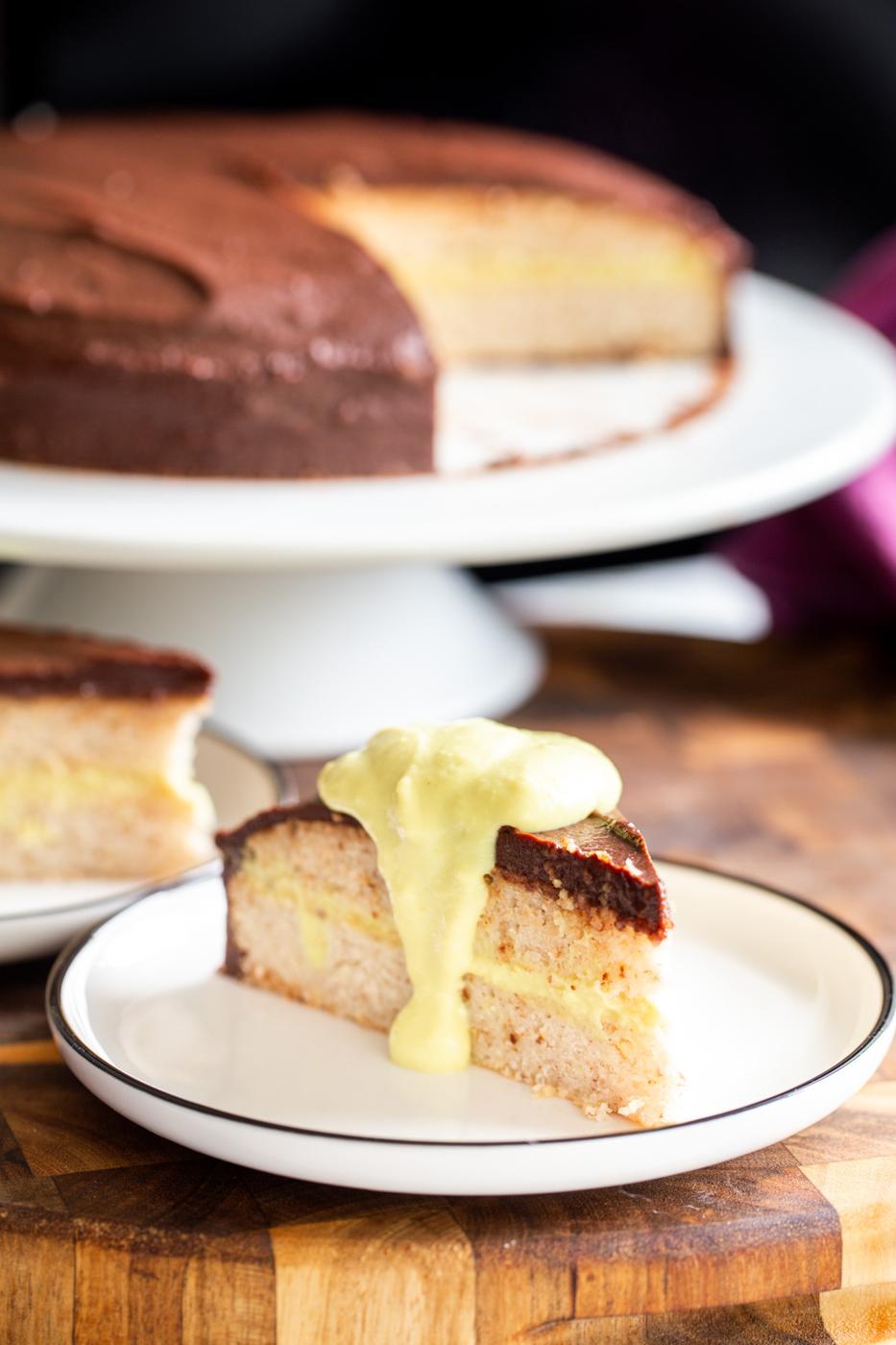 a slice of vegan boston cream pie cake with the leftover cake on a cakestand in the background