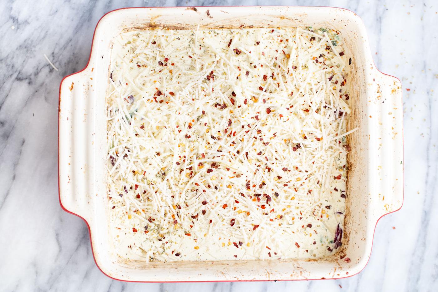 cheese shreds and pepper flakes being added over lasagna noodles