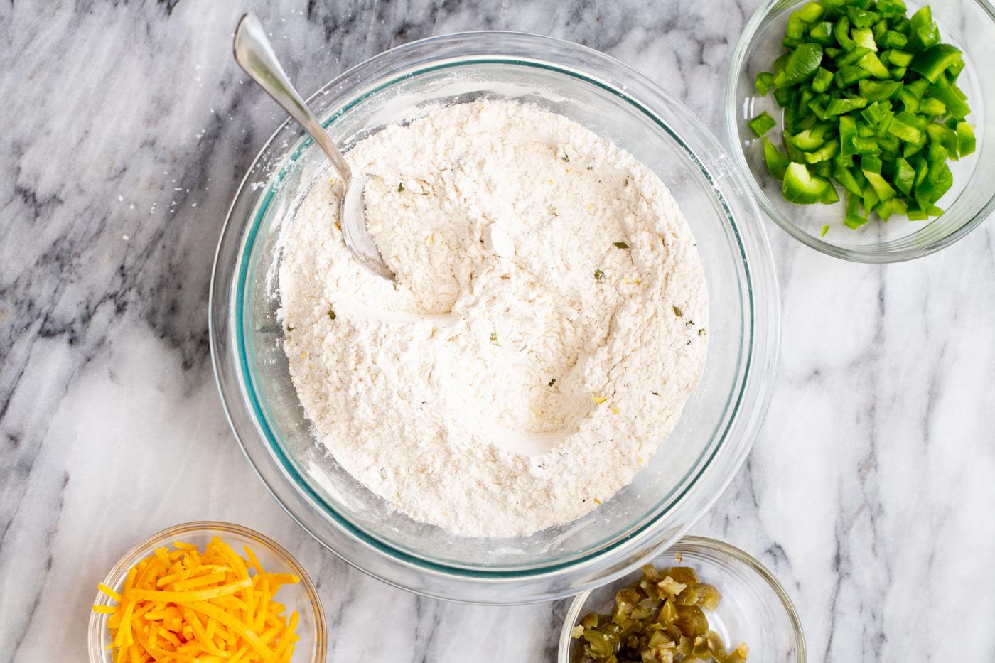 mixed dry ingredients in a glass bowl surrounded by ingredient dishes with jalapenos and cheddar