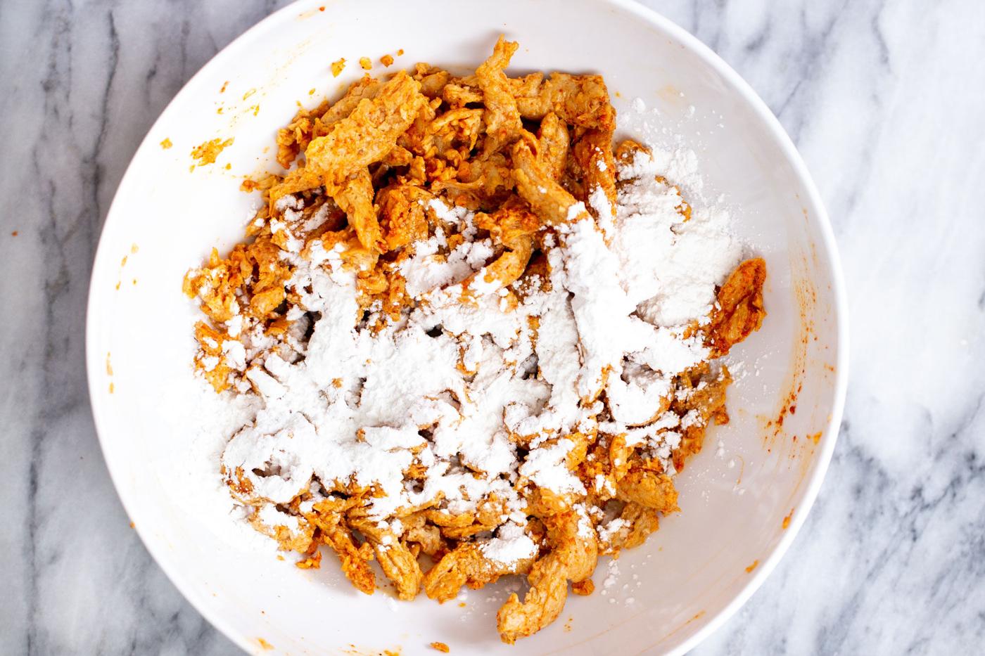 cornstarch being sprinkled over marinated soy curls in a white bowl