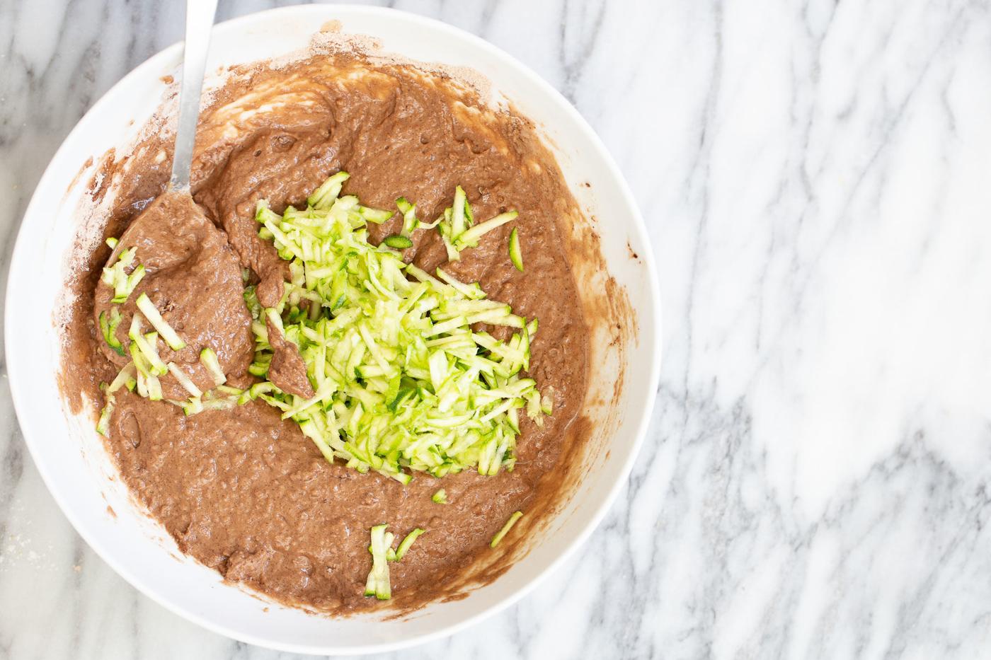 shredded zucchini being added to chocolate cake batter in a white mixing bowl