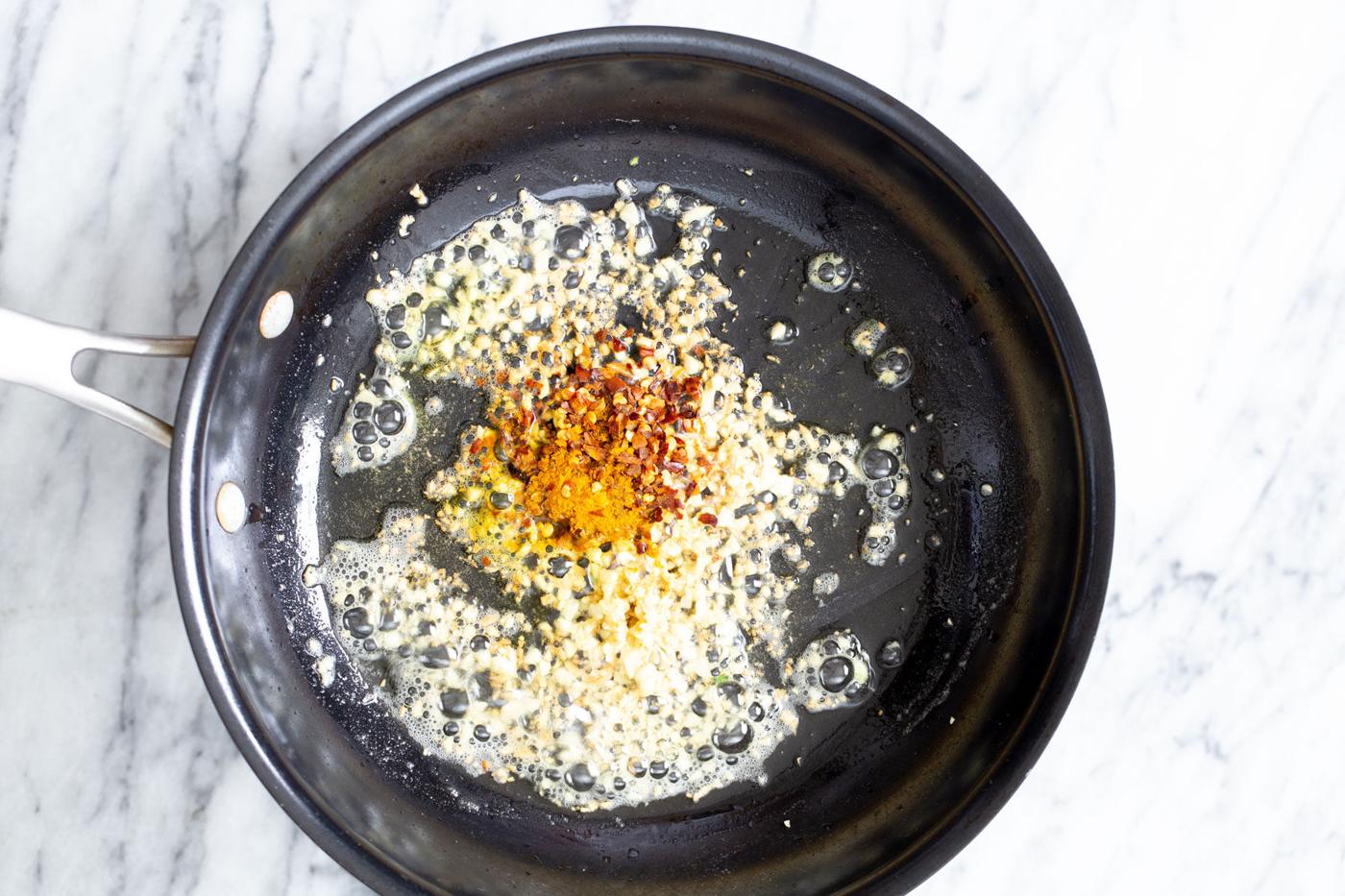 turmeric and pepper flakes being added to garlic in a frying pan