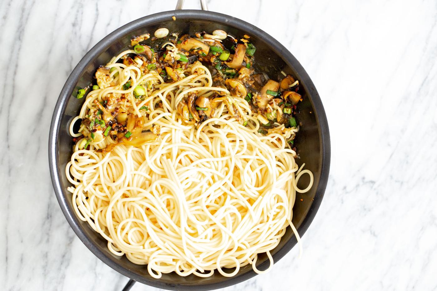 pasta being added to sauteed mushrooms, garlic and scallions in a pan to make garlic pasta