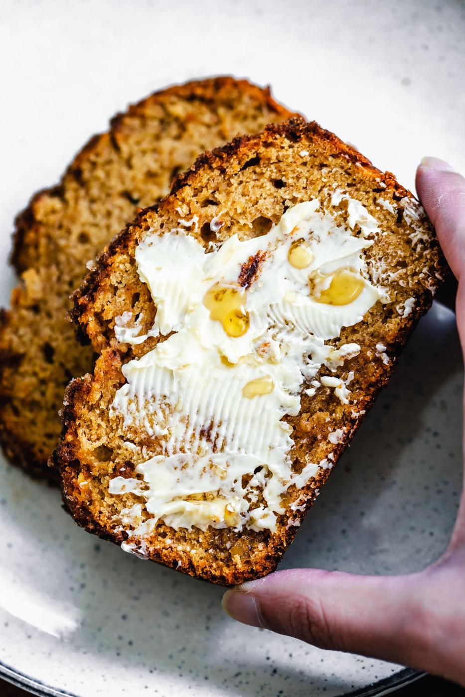 a hand reaching for a buttered slice of vegan gingerbread banana bread