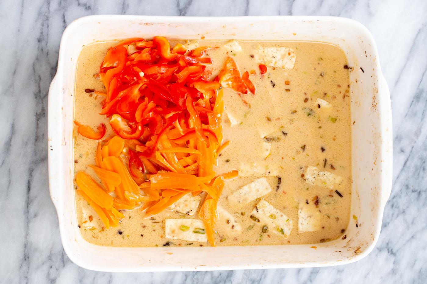 veggies being added to a casserole dish with tofu and peanut sauce