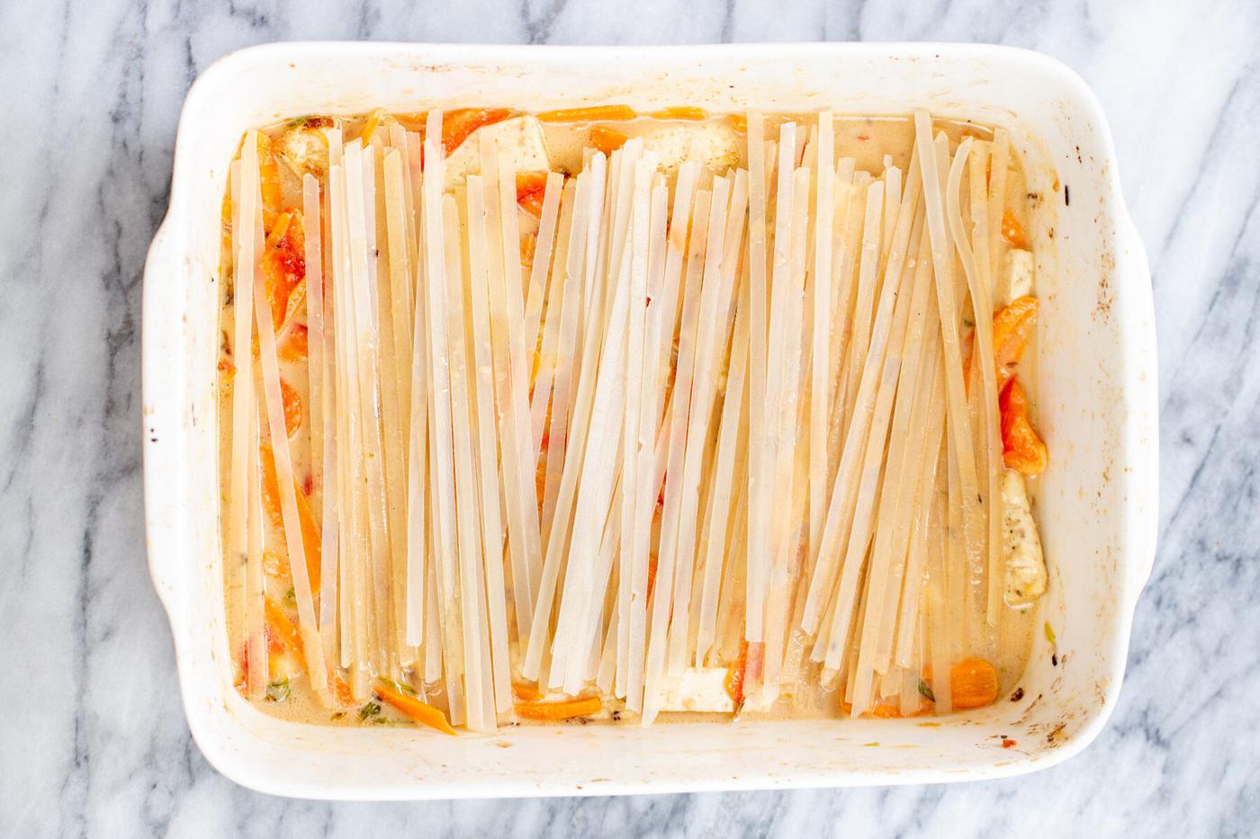 raw rice noodles being added to veggies and tofu in a white casserole dish