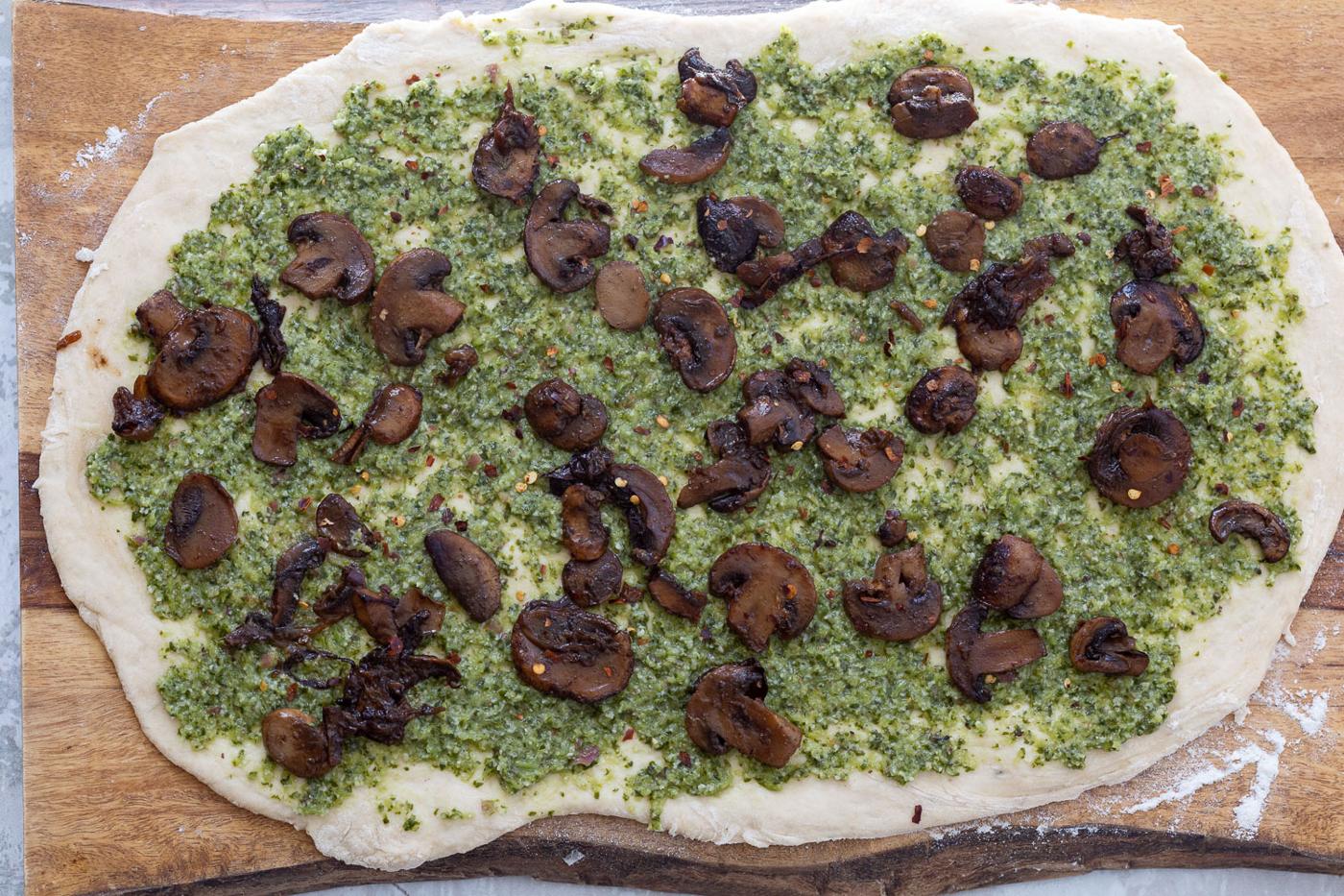 sauteed mushrooms being sprinkled onto rolled out pizza dough spread with pesto