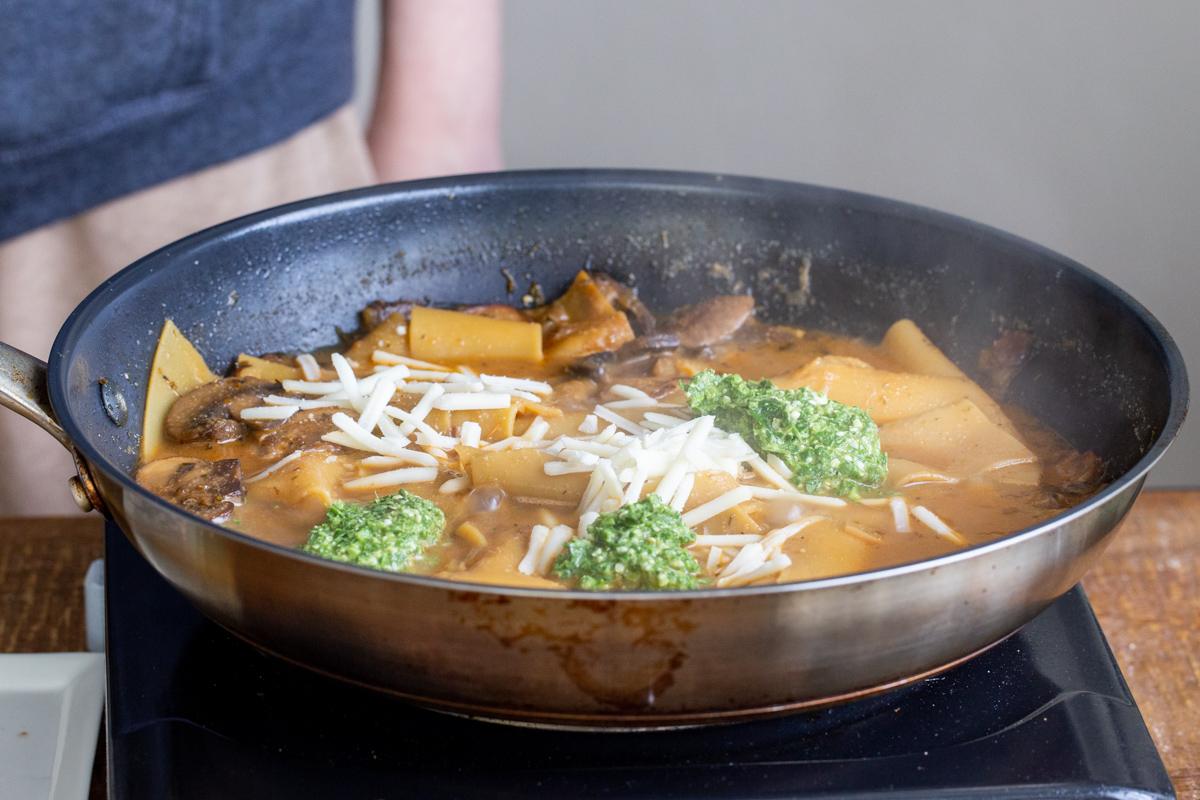 lasagna pasta simmering in a skillet topped with dollops of pesto and vegan cheese