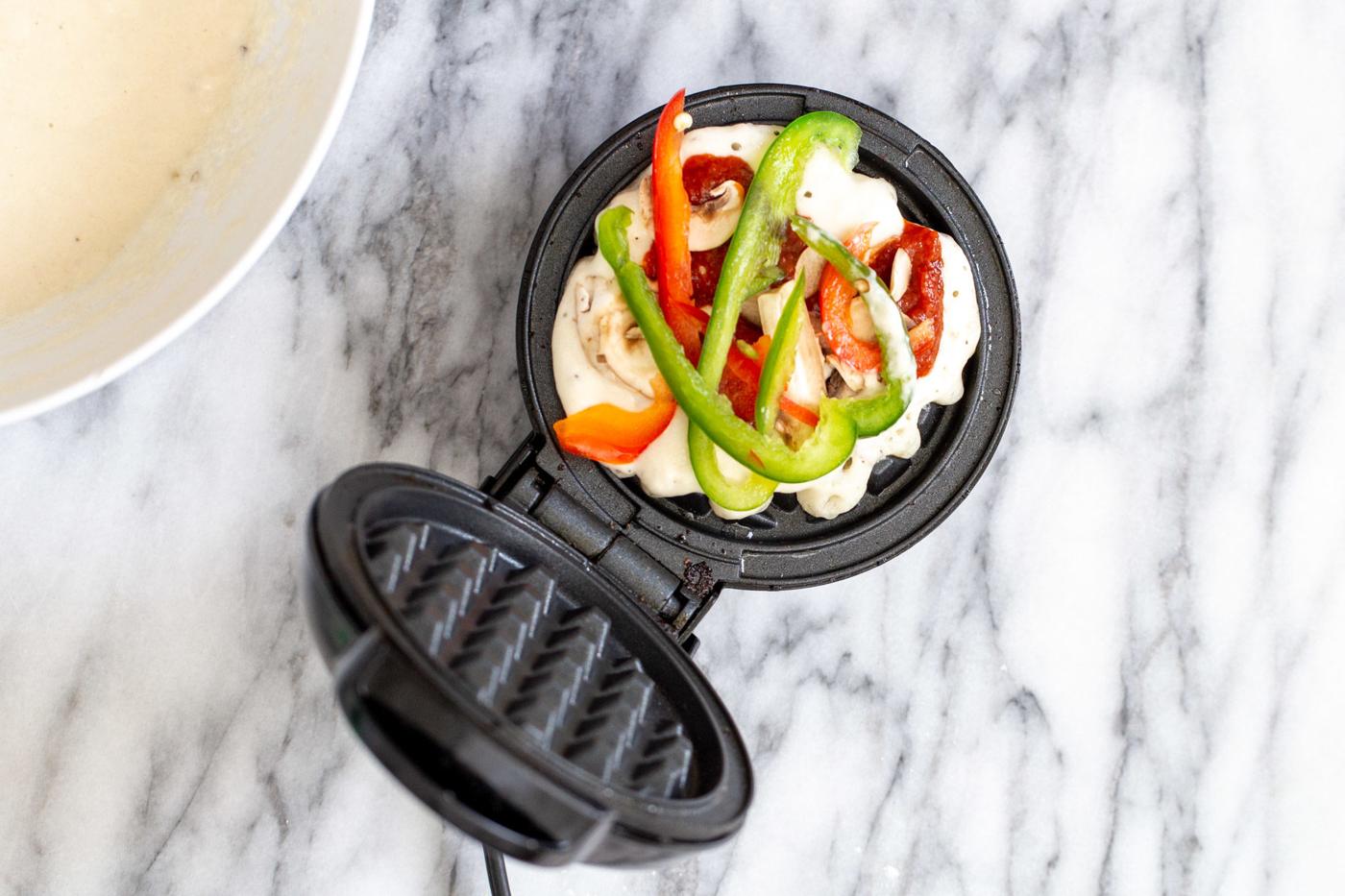 vegan pizza waffles batter in a waffle iron being topped with veggies