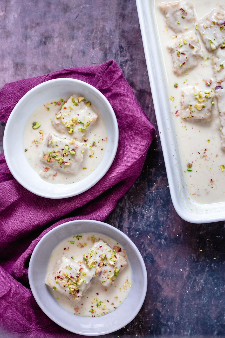 two servings of two plates of vegan rasmalai cake in milk sauce sprinkled with pistachios