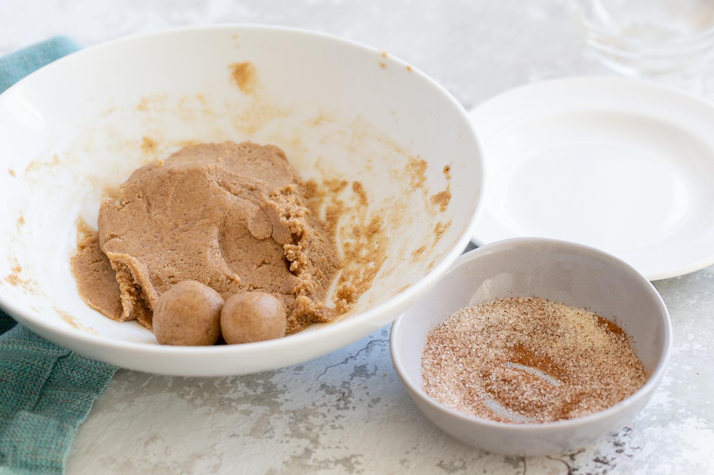 snickerdoodle bites being rolled in a bowl with some cinnamon sugar for coating on the side