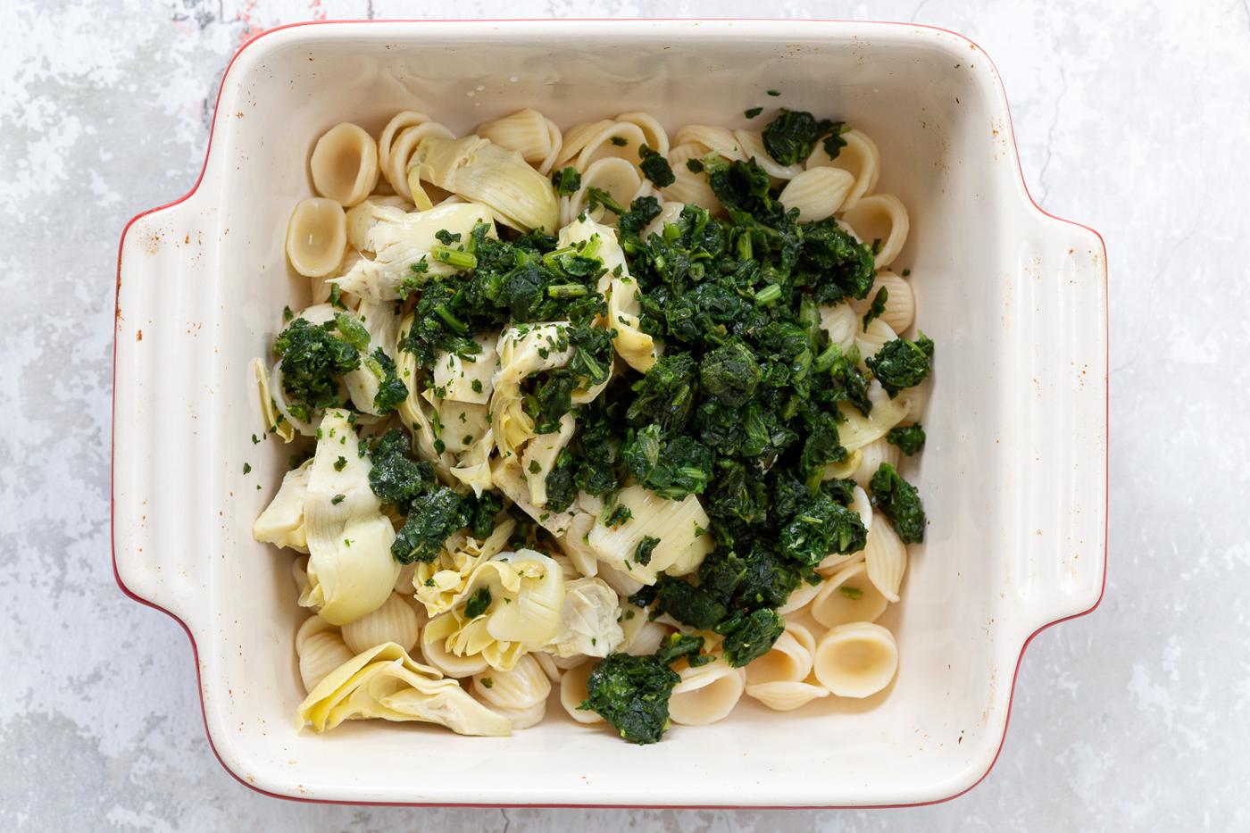overhead shot of a casserole dish with cooked shell pasta, spinach, and chopped artichokes