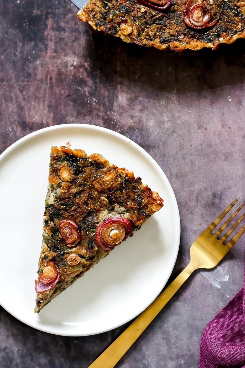 a slice of vegan spinach mushroom tart on a white plate with a golden fork on the side
