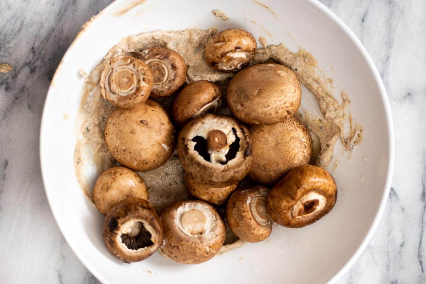 whole cremini mushrooms being tossed with marinade in a white bowl