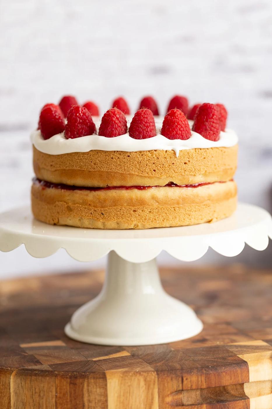 a vegan white cake topped with coconut whip and raspberries on a white cake stand