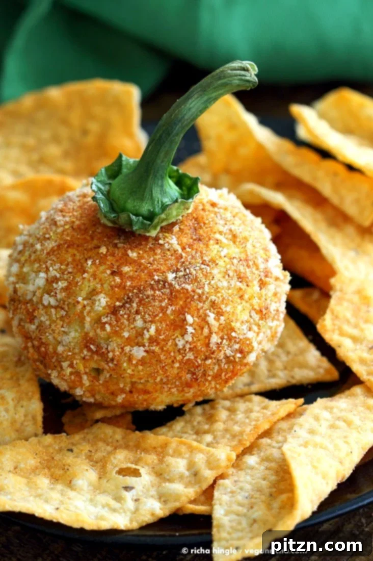 A festive Vegan Cheese Ball shaped like a pumpkin, garnished with a pretzel stem and surrounded by crackers. Perfect for Halloween, this nut-free, dairy-free appetizer is made with chickpea flour.