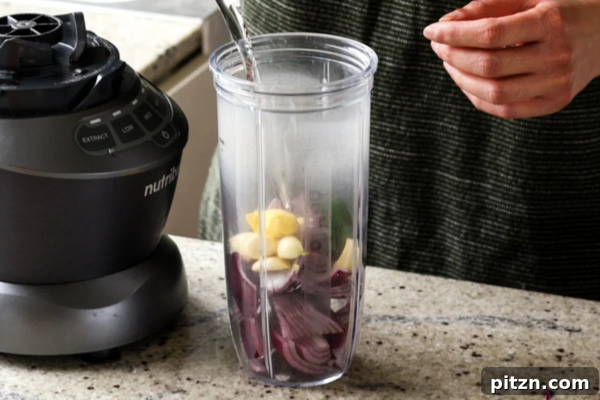 Adding water and aromatic ingredients like onion, garlic, ginger, and green chili to a blender jar for the masala paste.