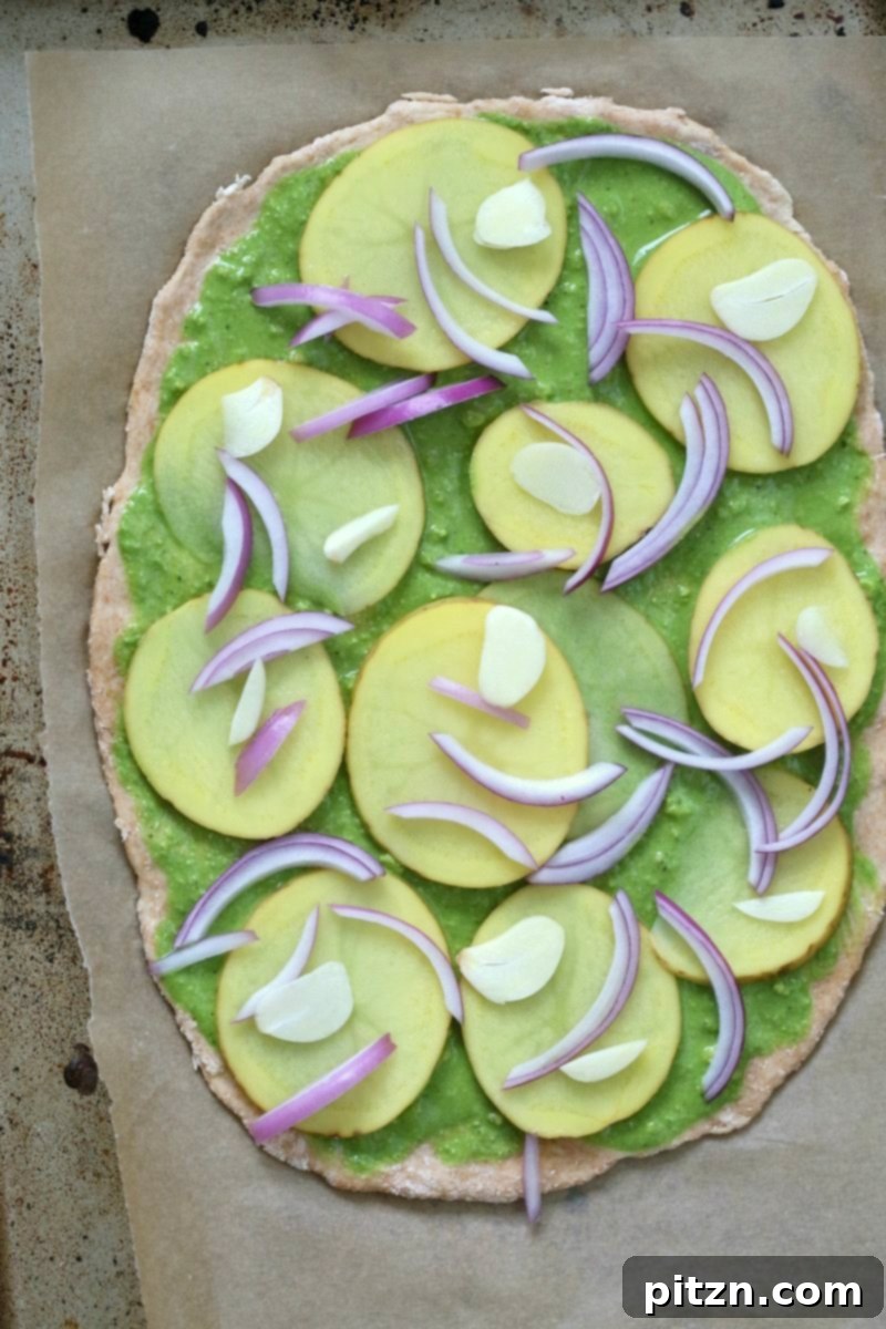 Vegan Basil Spinach Potato Pesto Pizza 4 Assembling the Potato Pesto Pizza, showing thinly sliced potatoes, red onion, and garlic layered over the pesto on a rustic thin crust.